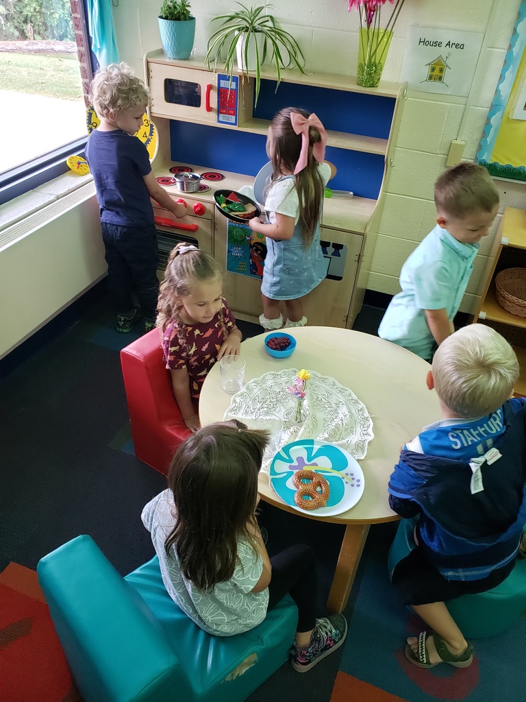 six children in a house area of a classroom.  some children are sitting around a table while two are at the kitchen getting food.