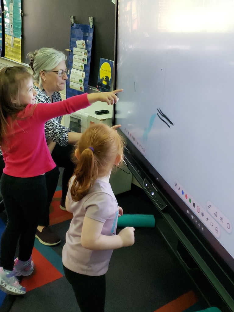 two children looking at a smart board with a teacher.  one child is pointing to the board.