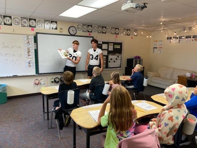 2 high school boys in white jerseys read a book to classroom of students.