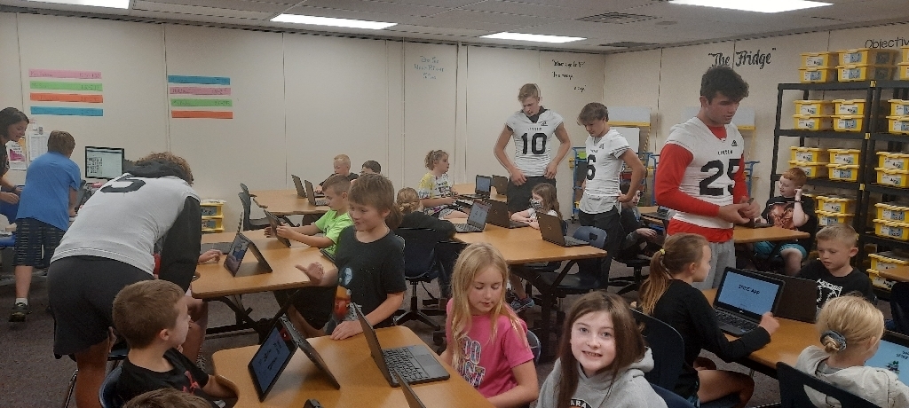 elementary aged students at desks with computers get help from high school student volunteers