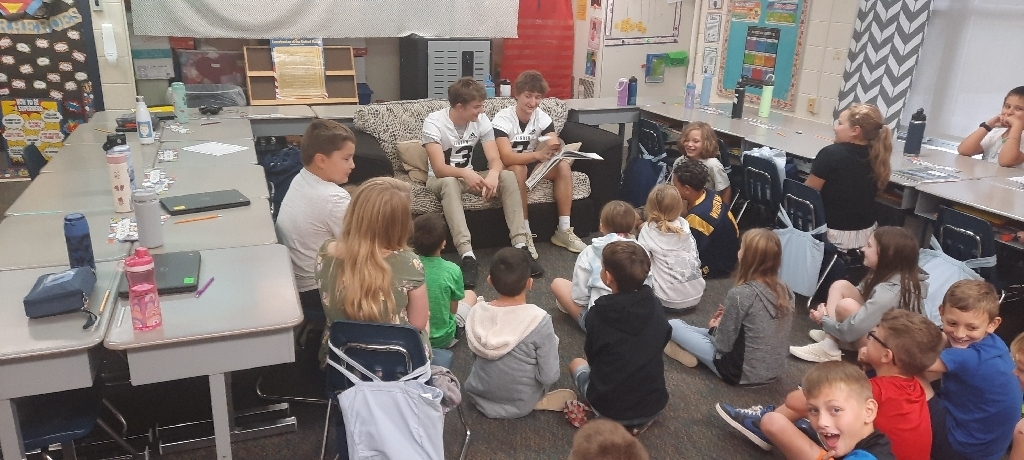 2 football players sit on couch in classroom holding a book, while elementary students sit on floor around them.