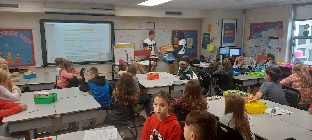 2 high school football players wearing white jerseys stand in front of class with a book, elementary students sit in desks facing the players.