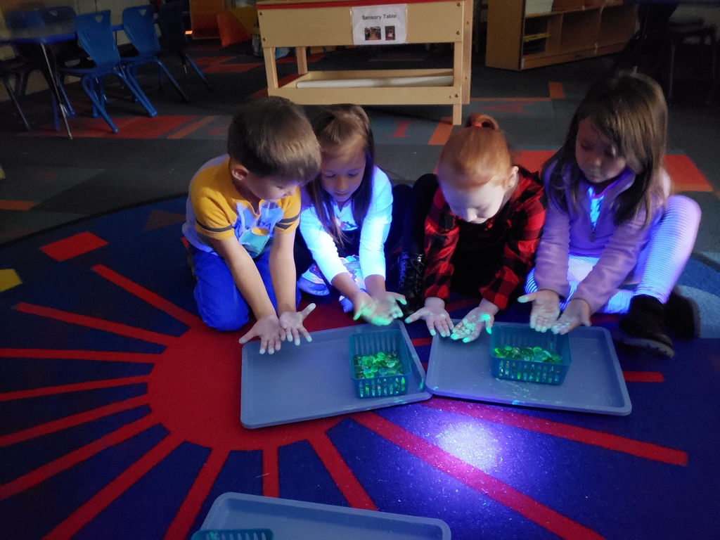 Four students holding out their hands and looking at the glowing stuff that is all over their fingertips and hands.  There is also two trays with a bin of glowing gems on each tray.