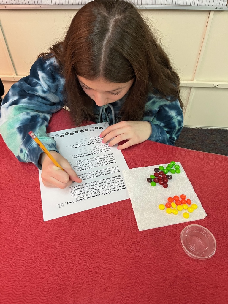 female student in blue patterned shirt working on math problem with Skittles separated into piles based on color.