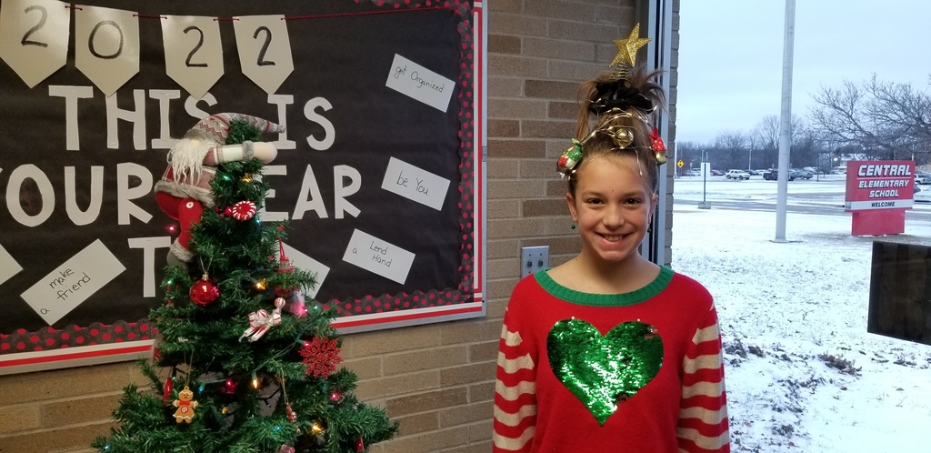 Girl standing next to Christmas tree, wearing red sweater with green sequin heart on it. Her brown hair is sticking up with a gold star on top and lights and ornaments wound up in it.