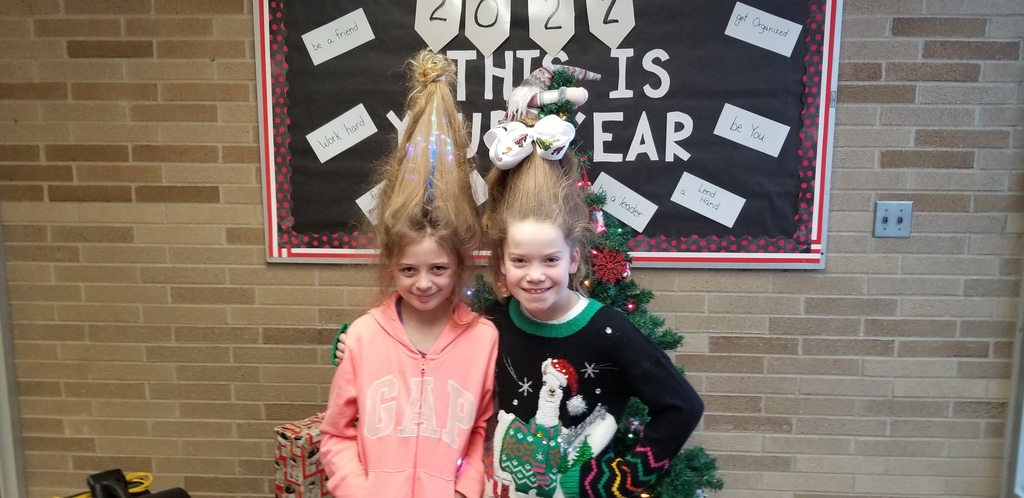 2 female students standing in front of Christmas tree with hair sticking up. Girl on left has blue lights mixed in her hair. Girl on right has white presents attached to hair. 