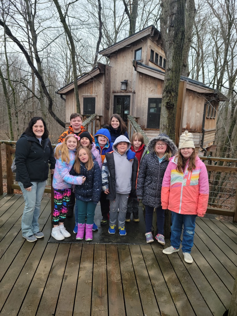 Group of elementary students in winter coats stand on deck in front of building in woods.