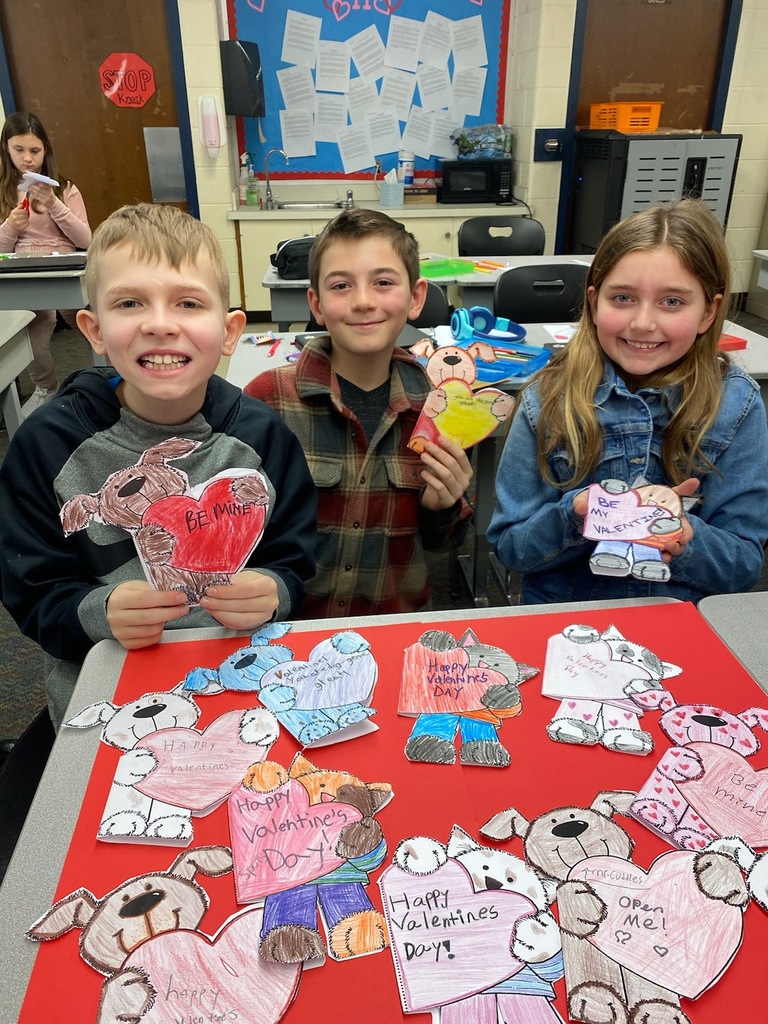 2 male and 1 female elementary aged students sitting in front of several hand made cards with hearts on them.