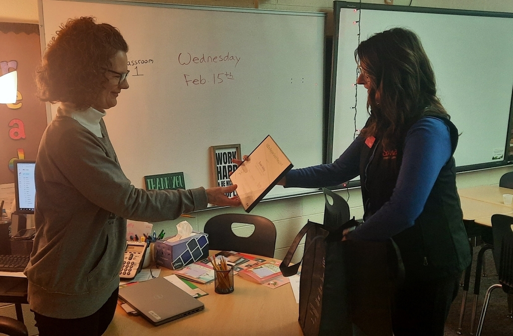 2 women standing near a desk. One is passing a certificate to the other.