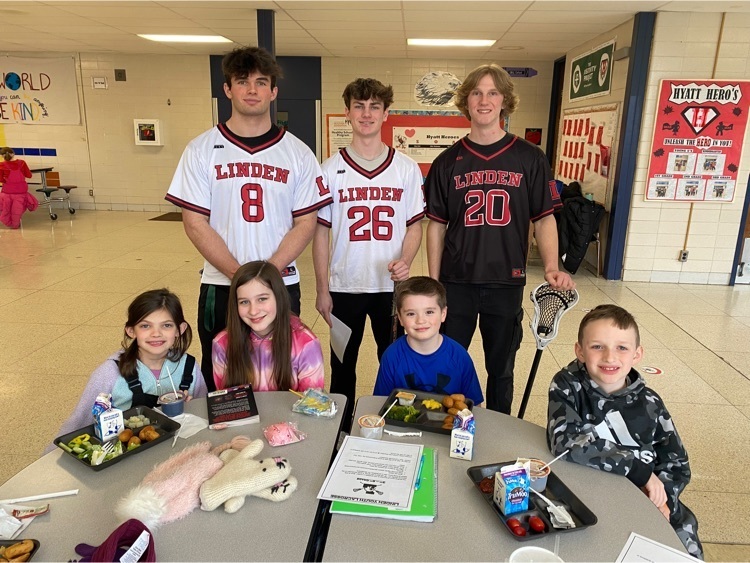 Three high school boys wearing the school lacrosse jerseys posing with third grade students in the cafeteria.