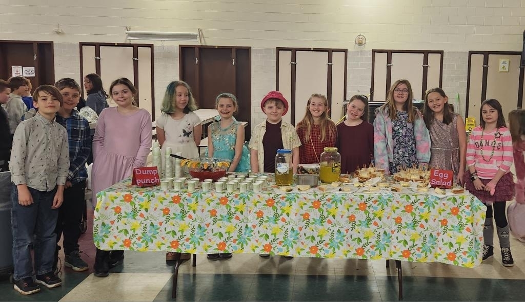 several students dressed up in fancy part attire standing in front of a table with a flower patterned table cloth that is full of food.