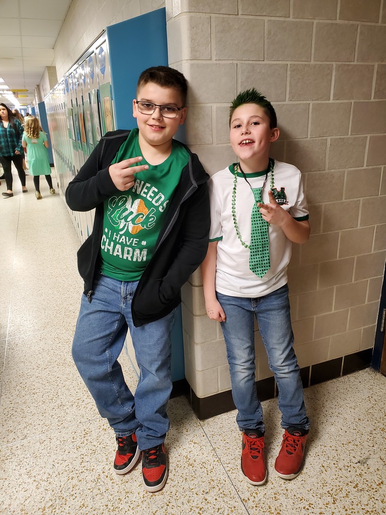 2 students, 1 wearing a green shirt, another with a white shirt and green tie standing next to wall.