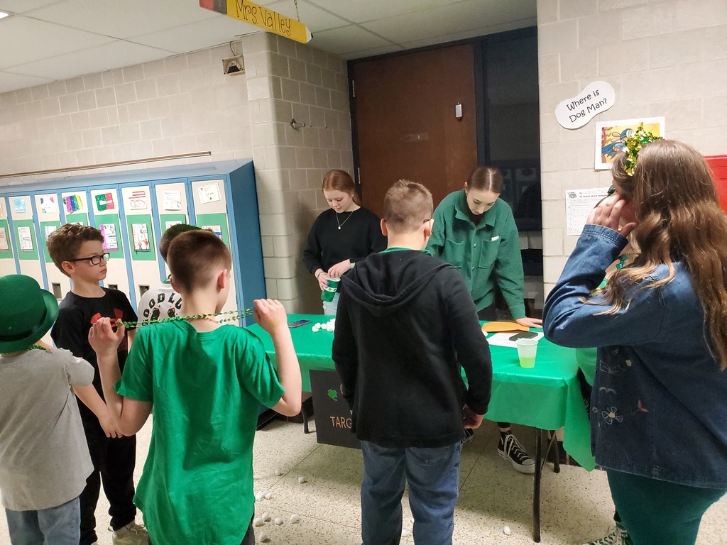 students in green surrounding a table with green table cloth playing a game facilitated by 2 adults wearing green.