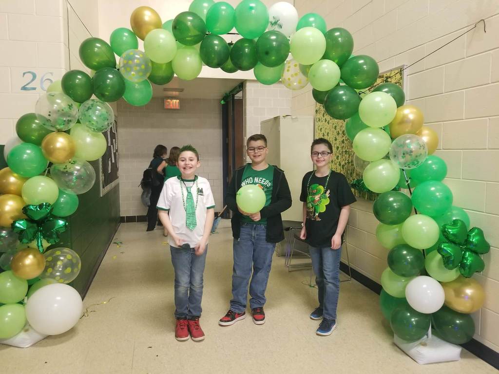 3 students wearing various green clothing standing under an arch of balloons that are several shades of green, along with white, gold and clear.