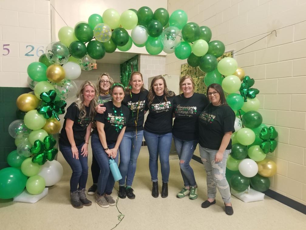 Group of parents wearing black shirts standing under a balloon arch full of green, white, gold and clear balloons.