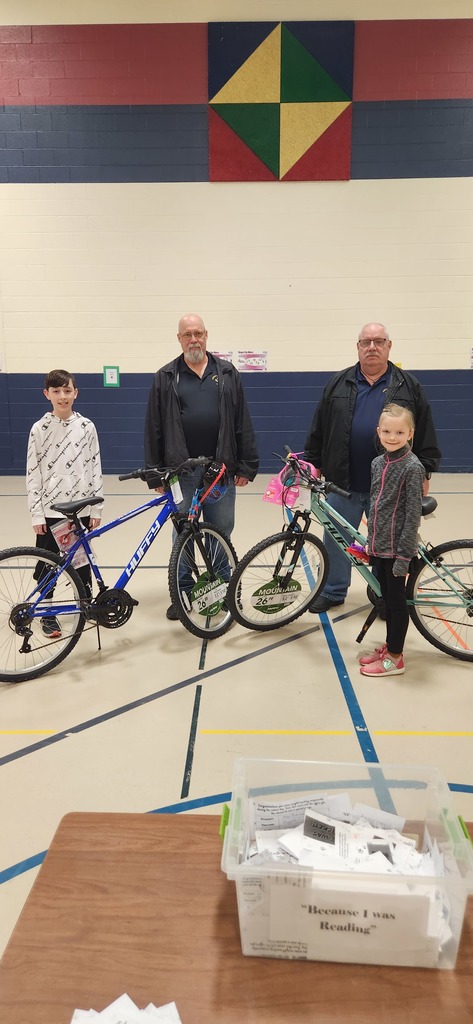 2 adult males in black sweatshirts stand next to a male and female elementary student and their new blue and teal bikes. 