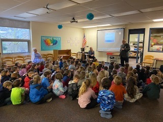Second grade children sitting on the floor of a library listening to a standing adult about the history of Linden. A teacher is sitting in a blue dress to their left and another teacher is sitting near the adult speaker.
