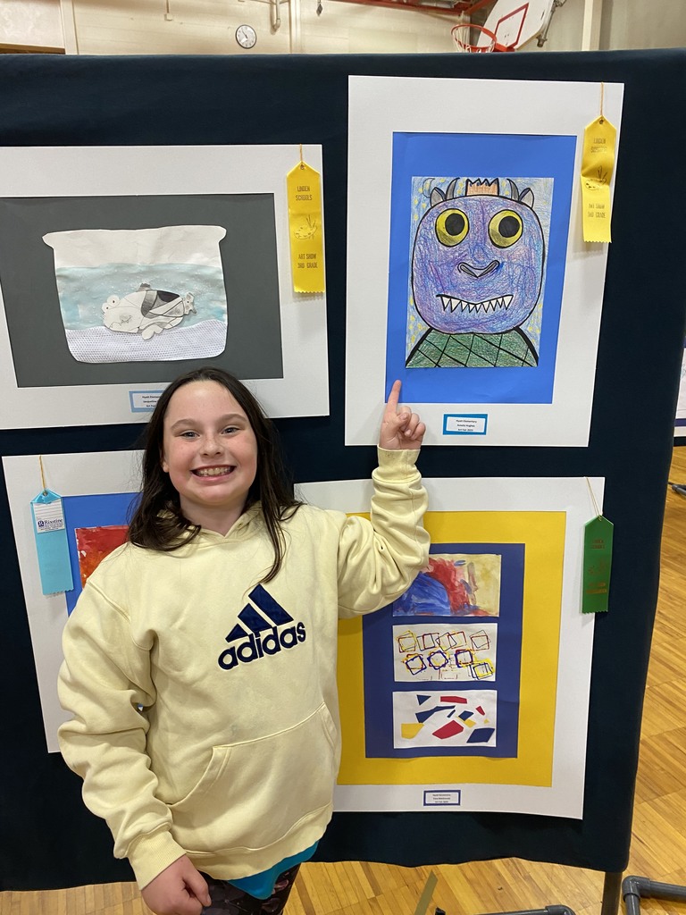 A student smiling in a yellow, adidas sweatshirt, pointing at her framed artwork.