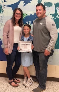 Mother in a pink sweater and white shirt is standing with her husband in a grey long sleeved shirt with their daughter who is wearing a blue dress and holding a student of the month certificate.